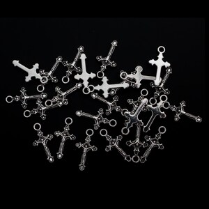 May include: A collection of silver-toned metal cross charms featuring intricate detailing and a small circular loop at the top for attachment. These small religious-themed pendants are scattered across the frame, showcasing a metallic finish.