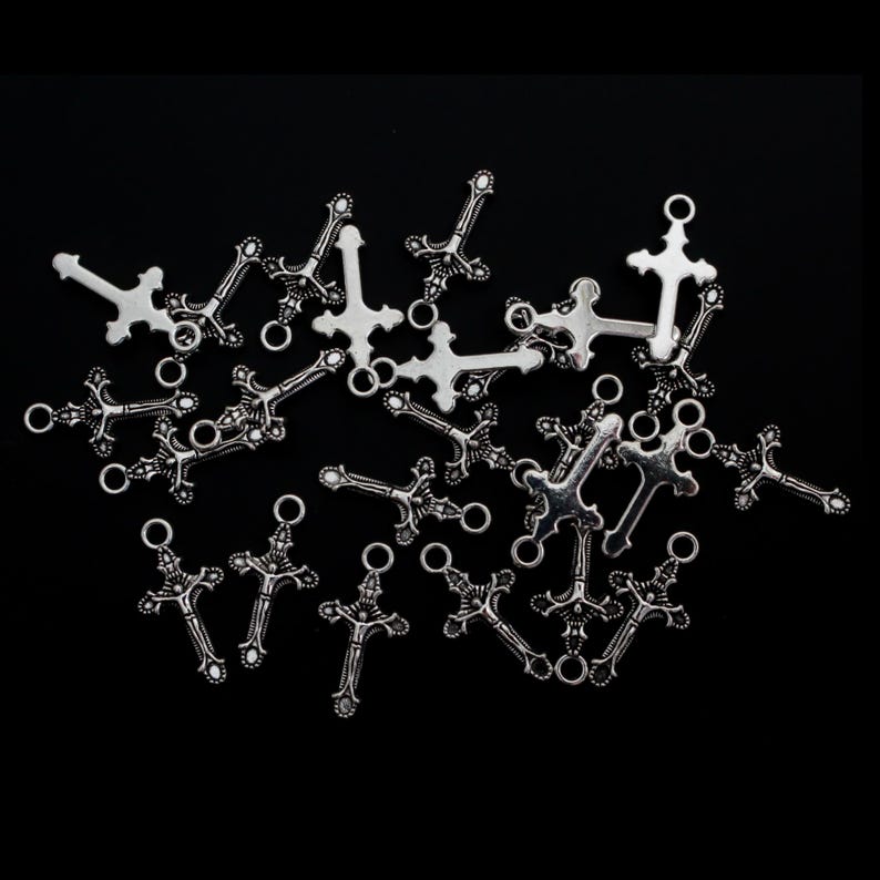 May include: A collection of silver-toned metal cross charms featuring intricate detailing and a small circular loop at the top for attachment. These small religious-themed pendants are scattered across the frame, showcasing a metallic finish.