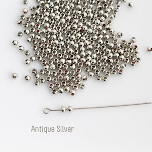May include: A collection of small, round, metallic beads with an antique silver finish. The beads are scattered in a cluster, with two beads threaded onto a thin metal headpin featuring a loop at one end. The text Antique Silver is visible.