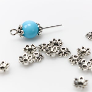 Puede incluir: Una colecci&oacute;n de tapas de cuentas de metal plateado y una cuenta azul claro con una tapa y un alfiler de metal plateado. Las tapas de las cuentas tienen forma de peque&ntilde;as flores y est&aacute;n dispersas sobre una superficie blanca.