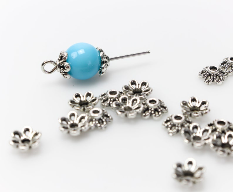 Puede incluir: Una colecci&oacute;n de tapas de cuentas de metal plateado y una cuenta azul claro con una tapa y un alfiler de metal plateado. Las tapas de las cuentas tienen forma de peque&ntilde;as flores y est&aacute;n dispersas sobre una superficie blanca.