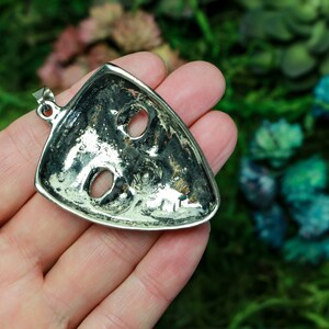 May include: Silver pendant in the shape of a mask with two eye holes. The pendant is on a hand against a blurred background of green and blue.