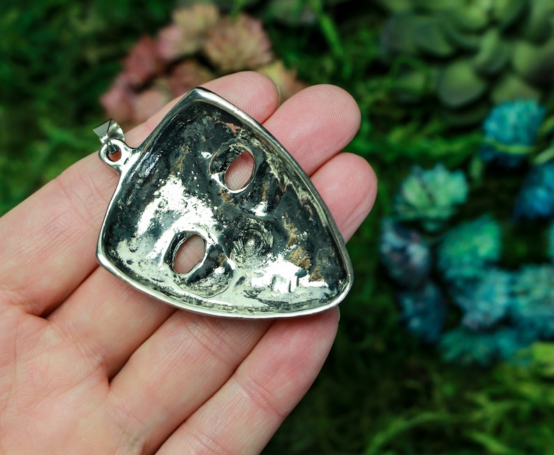 May include: Silver pendant in the shape of a mask with two eye holes. The pendant is on a hand against a blurred background of green and blue.