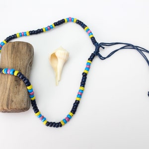 May include: A blue and black beaded necklace with pink, yellow, and blue accents. The necklace has a simple knot closure and is displayed on a piece of driftwood with a seashell.