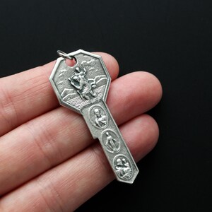 May include: A silver key-shaped charm with a religious design. The charm features a figure of a saint on the top and four smaller figures of saints on the bottom.