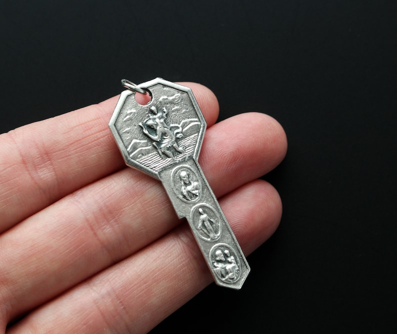 May include: A silver key-shaped charm with a religious design. The charm features a figure of a saint on the top and four smaller figures of saints on the bottom.