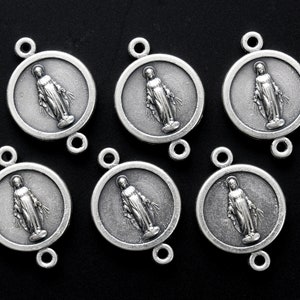 May include: Six silver-toned metal charms featuring a depiction of the Virgin Mary. Each charm is round with a raised image of the Virgin Mary in the centre. The charms have two loops at the top and bottom for attaching to a necklace or bracelet.