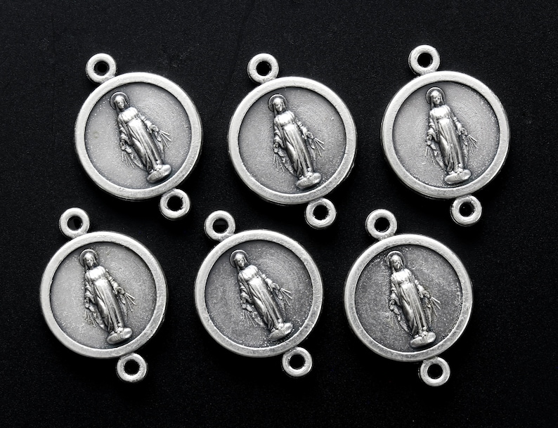 May include: Six silver-toned metal charms featuring a depiction of the Virgin Mary. Each charm is round with a raised image of the Virgin Mary in the centre. The charms have two loops at the top and bottom for attaching to a necklace or bracelet.