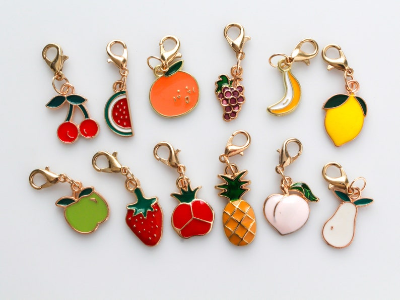 Pu&ograve; includere: Un set di 10 ciondoli di frutta dorati con dettagli smaltati. I ciondoli includono una ciliegia, una fetta di anguria, un'arancia, dell'uva, una banana, un limone, una mela, una fragola, un pomodoro, un ananas e una pesca.