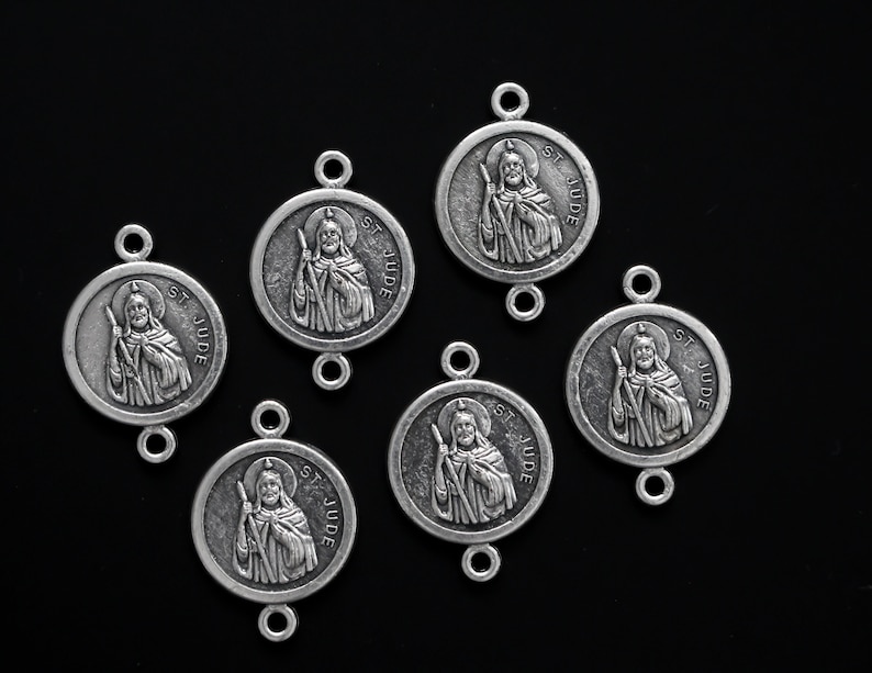 May include: Six silver-toned metal charms featuring a depiction of Saint Jude Thaddeus. Each charm has a loop on the top and bottom for attaching to a chain or bracelet. The charms are circular and have the words "St Jude" inscribed around the edge.