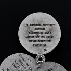 May include: Close-up of a silver religious pendant with multiple circular charms. The top charm reads "THE LUMINOUS MYSTERIES" and lists "BAPTISM", "WEDDINGS AT CANA", "SERMON ON THE MOUNT", "TRANSFIGURATION", and "EUCHARIST".