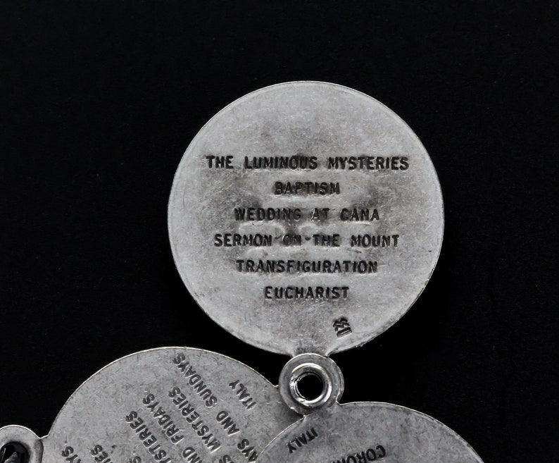 May include: Close-up of a silver religious pendant with multiple circular charms. The top charm reads "THE LUMINOUS MYSTERIES" and lists "BAPTISM", "WEDDINGS AT CANA", "SERMON ON THE MOUNT", "TRANSFIGURATION", and "EUCHARIST".