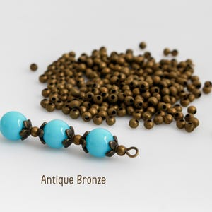 May include: A collection of small, round, antique bronze metal spacer beads scattered alongside a string of three smooth, bright turquoise beads, each accented by two bronze flower-shaped bead caps and a small metal loop at the end. Text reads Antique Bronze.