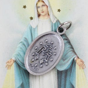 May include: Silver oval charm with a depiction of the Virgin Mary and stars. The charm is hanging in front of a vintage image of the Virgin Mary.