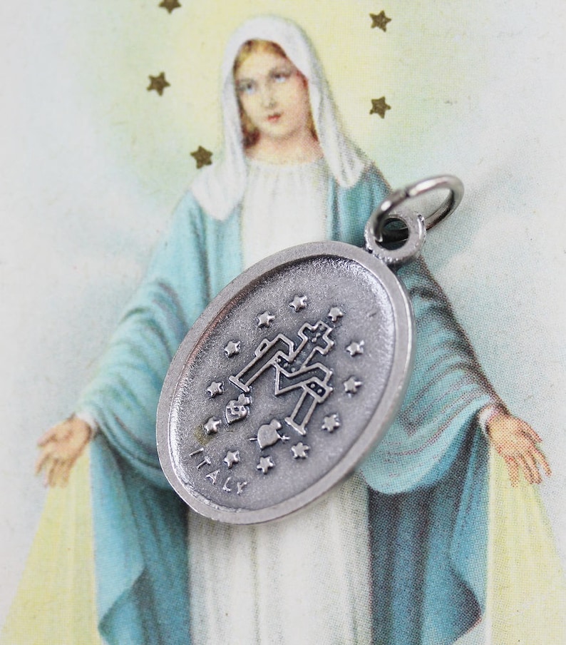 May include: Silver oval charm with a depiction of the Virgin Mary and stars. The charm is hanging in front of a vintage image of the Virgin Mary.