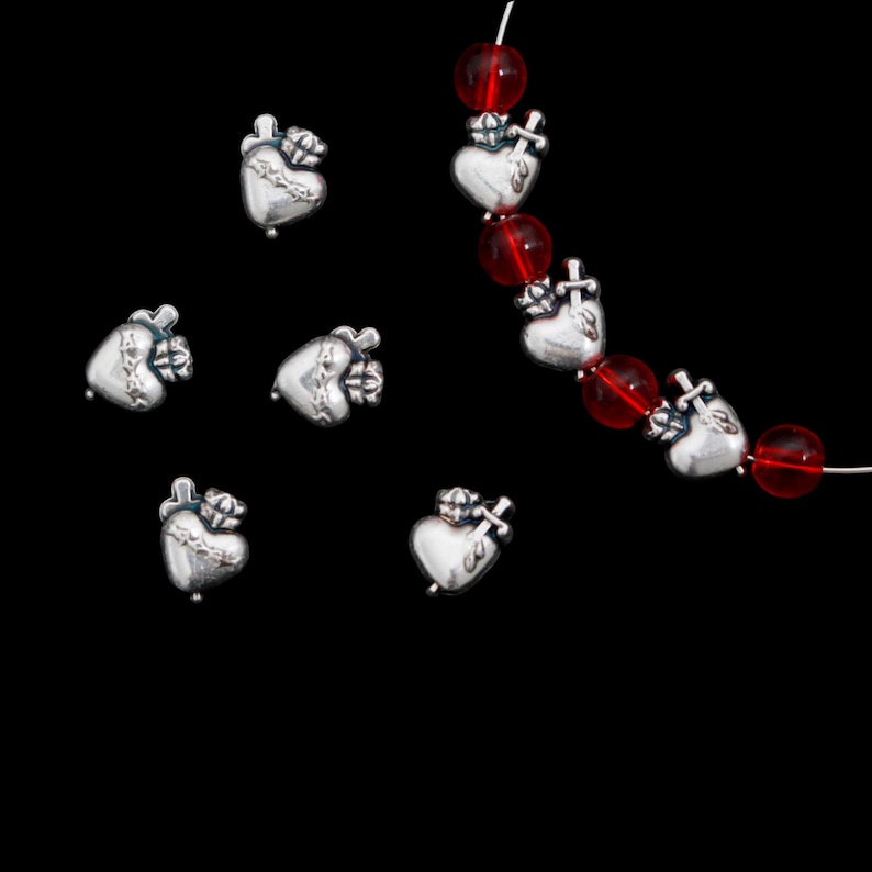 May include: Silver-toned heart-shaped charms with a crown and cross design. Some charms are strung with red beads on a white string. The charms are scattered against a black background, creating a striking contrast.