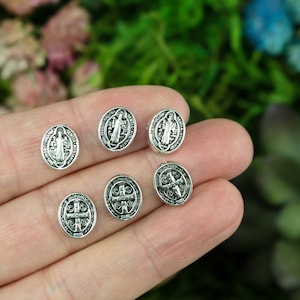 May include: Six silver-toned metal charms featuring a religious icon. The charms are oval-shaped and have a raised design.