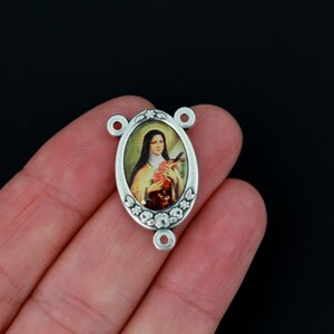 May include: Silver oval-shaped charm with a floral design and a picture of Saint Th&eacute;r&egrave;se of Lisieux holding a rose.