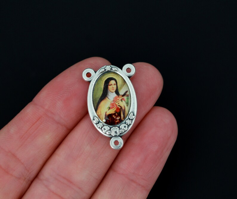 May include: Silver oval-shaped charm with a floral design and a picture of Saint Th&eacute;r&egrave;se of Lisieux holding a rose.