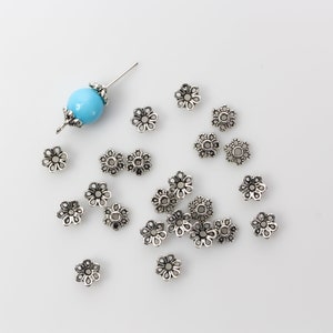 Puede incluir: Un conjunto de tapas de cuentas plateadas en forma de flor esparcidas sobre una superficie blanca. Tambi&eacute;n hay una cuenta azul claro con detalles plateados y un alfiler. Estas tapas de cuentas decorativas son ideales para la fabricaci&oacute;n de joyas.