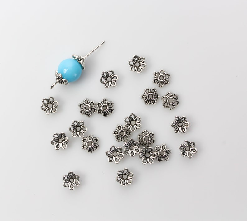 Puede incluir: Un conjunto de tapas de cuentas plateadas en forma de flor esparcidas sobre una superficie blanca. Tambi&eacute;n hay una cuenta azul claro con detalles plateados y un alfiler. Estas tapas de cuentas decorativas son ideales para la fabricaci&oacute;n de joyas.