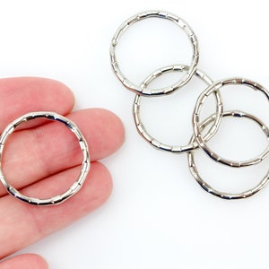 May include: A collection of silver-colored metal rings, each with a textured, segmented design. One ring is held by a hand, while the others are interlocked, creating a decorative pattern. The rings are on a white background.