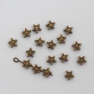 May include: A collection of small, antique bronze-colored metal star charms. Each star features a raised outline and a recessed center. One charm is attached to a thin metal wire loop, while the others are scattered individually across the frame.