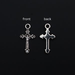 May include: Two silver-toned metal cross charms, each featuring a circular jump ring at the top. The left charm displays a detailed crucifix figure on the front, while the right charm shows the smooth, plain back side of the same cross design.