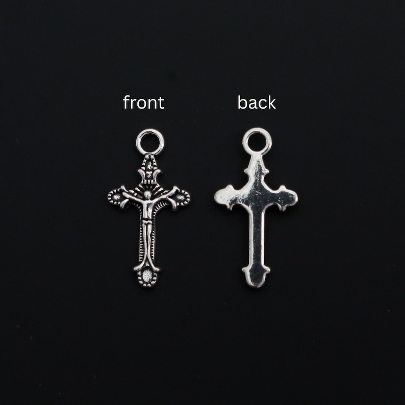 May include: Two silver-toned metal cross charms, each featuring a circular jump ring at the top. The left charm displays a detailed crucifix figure on the front, while the right charm shows the smooth, plain back side of the same cross design.