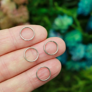 May include: Four small, silver hoop earrings are shown on a person's fingers. The earrings are circular and appear to be made of metal. The background is a soft focus of green and blue.