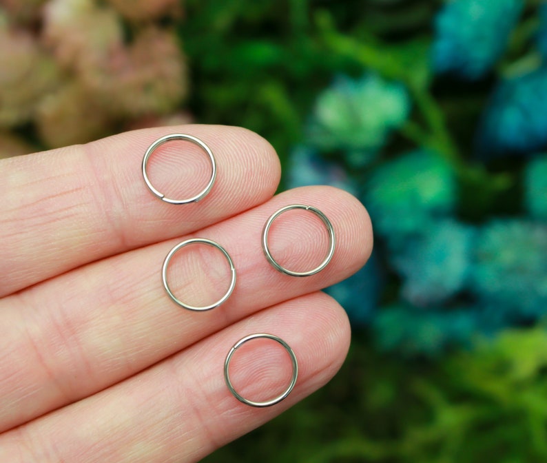 May include: Four small, silver-colored hoop earrings are displayed on a person's fingers. The earrings are circular and appear to be made of metal. The background is a soft focus of green and blue.
