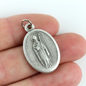 May include: Silver oval pendant with a depiction of Saint Augustine holding a book and a staff. The pendant has the text "St. Augustine" inscribed on it.