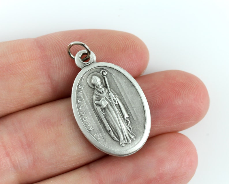 May include: Silver oval pendant with a depiction of Saint Augustine holding a book and a staff. The pendant has the text "St. Augustine" inscribed on it.
