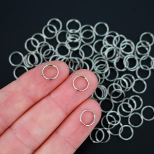 May include: A close-up of a hand holding three silver jump rings. The rings are small and circular, and they are arranged in a row on the fingers. There are many more silver jump rings scattered on a black surface behind the hand.