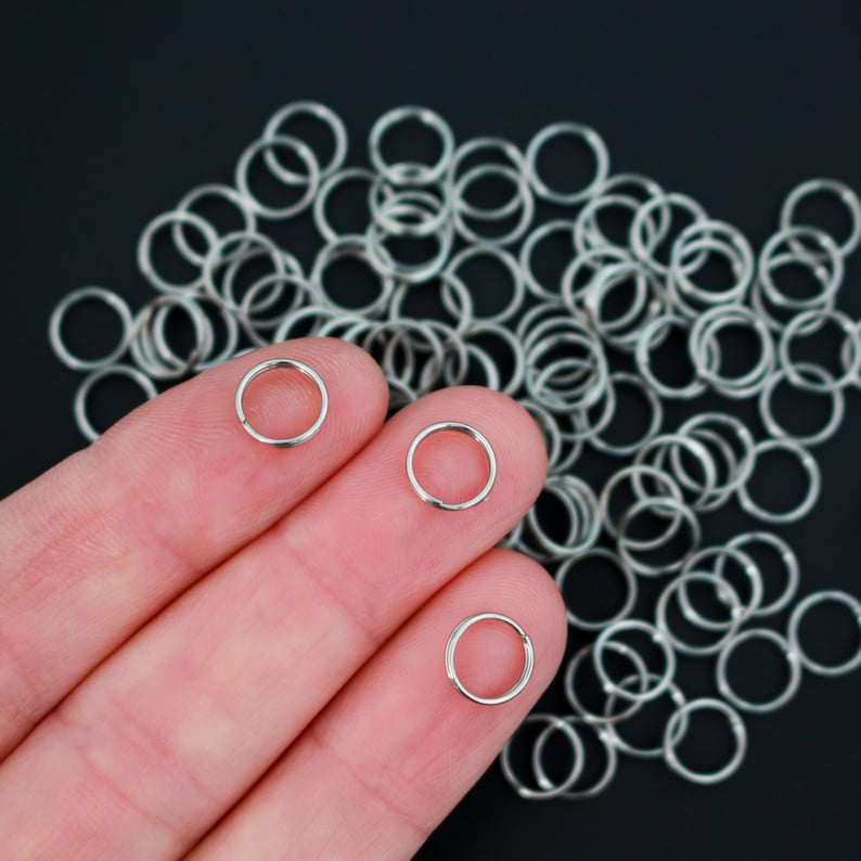 May include: A close-up of a hand holding three silver jump rings. The rings are small and circular, and they are arranged in a row on the fingers. There are many more silver jump rings scattered on a black surface behind the hand.