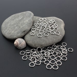 K&ouml;nnte beinhalten: Eine Ansammlung kleiner, kreisf&ouml;rmiger, silberfarbener Metall-Biegeringe, die auf einer dunklen Oberfl&auml;che verstreut sind. Einige Ringe liegen auf zwei glatten, grauen, ovalen Steinen, w&auml;hrend andere in einer Gruppe unterhalb liegen.