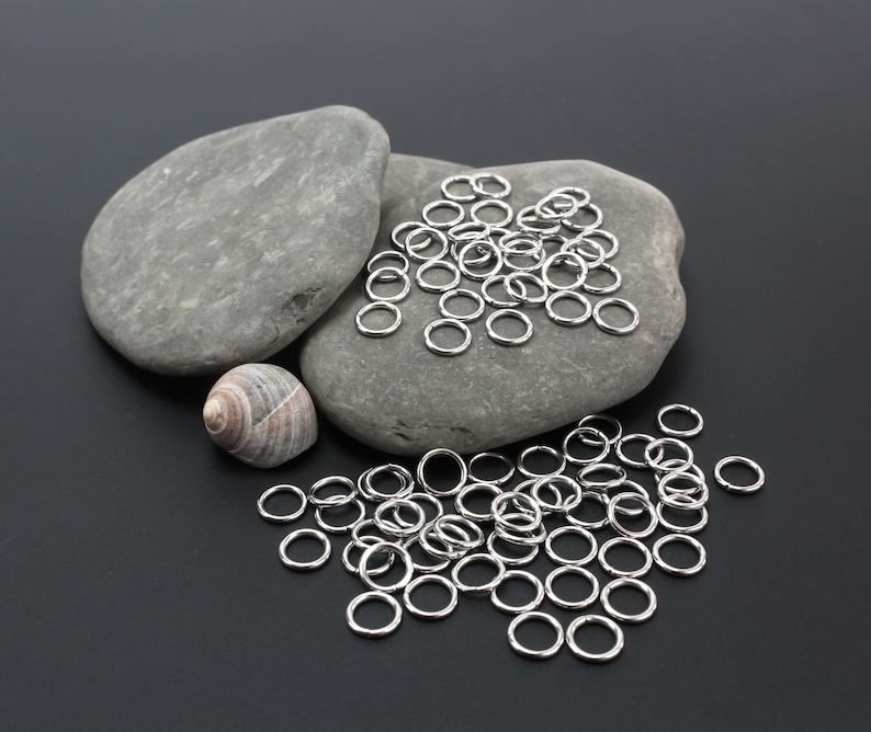 K&ouml;nnte beinhalten: Eine Ansammlung kleiner, kreisf&ouml;rmiger, silberfarbener Metall-Biegeringe, die auf einer dunklen Oberfl&auml;che verstreut sind. Einige Ringe liegen auf zwei glatten, grauen, ovalen Steinen, w&auml;hrend andere in einer Gruppe unterhalb liegen.