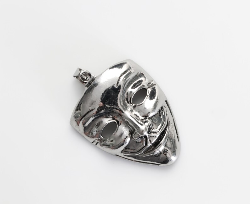 May include: A silver pendant in the shape of a mask with a mischievous grin and two black eyes. The mask is a popular symbol of anonymity and privacy.