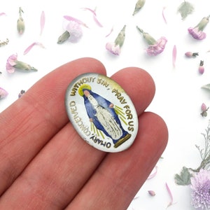 May include: A silver oval-shaped religious medal with a gold border and a blue and white image of the Virgin Mary. The medal has the text "OUR LADY CONCEIVED WITHOUT SIN, PRAY FOR US" around the border.