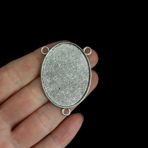 Puede incluir: Base de colgante ovalada plateada con tres pequeños bucles para sujetar a una cadena u otros componentes de joyería. El colgante tiene una superficie texturizada y se sostiene en una mano sobre un fondo negro.