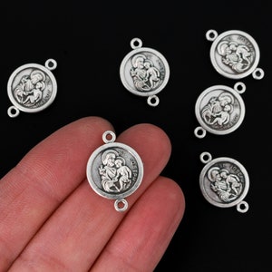 Op de afbeelding: Zilverkleurige St. Joseph connector bedels. Elke bedel toont een afbeelding van St. Joseph en een kind, met de woorden "ST. JOSEPH" rond de rand. De bedels hebben twee kleine lussen om aan andere sieradenonderdelen te bevestigen.
