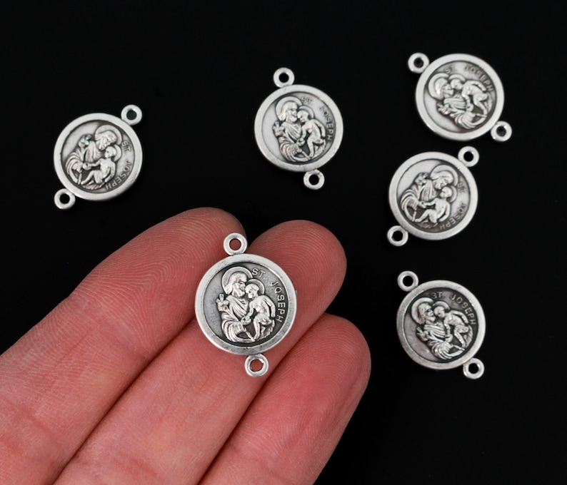 Op de afbeelding: Zilverkleurige St. Joseph connector bedels. Elke bedel toont een afbeelding van St. Joseph en een kind, met de woorden "ST. JOSEPH" rond de rand. De bedels hebben twee kleine lussen om aan andere sieradenonderdelen te bevestigen.