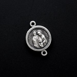 May include: A silver-toned religious pendant with the words "HOLY FAMILY ITALY" encircling a detailed relief of a family. The pendant has two small loops for attachment and is set against a black background.