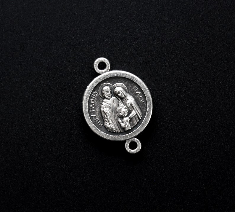 May include: A silver-toned religious pendant with the words "HOLY FAMILY ITALY" encircling a detailed relief of a family. The pendant has two small loops for attachment and is set against a black background.