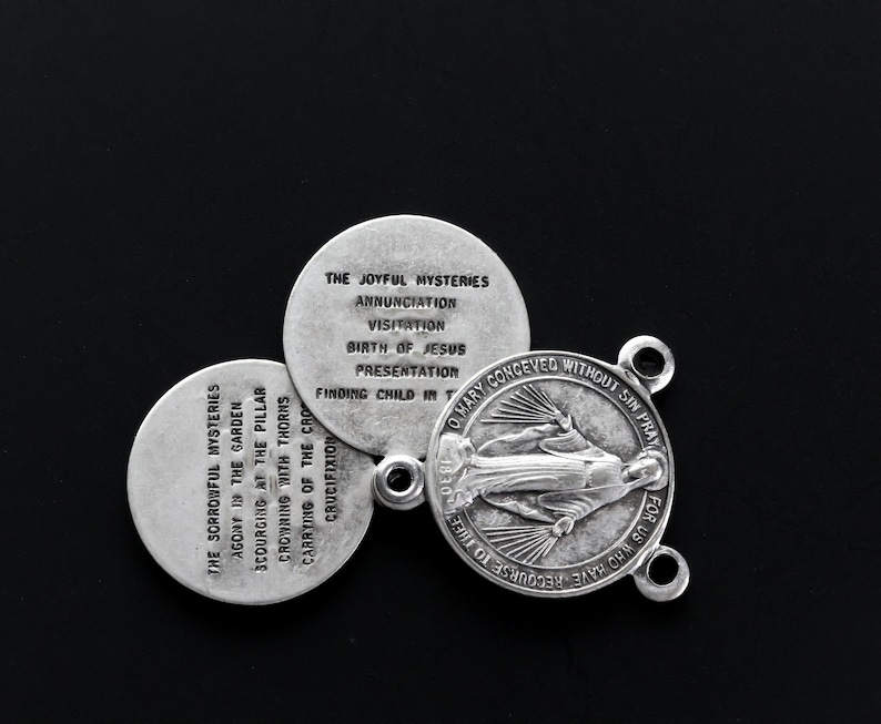 May include: A silver rosary centre and three round medallions. The centre features an image of the Virgin Mary and the text "O Mary conceived without sin, pray for us who have recourse to thee." The medallions list the Joyful and Sorrowful Mysteries.