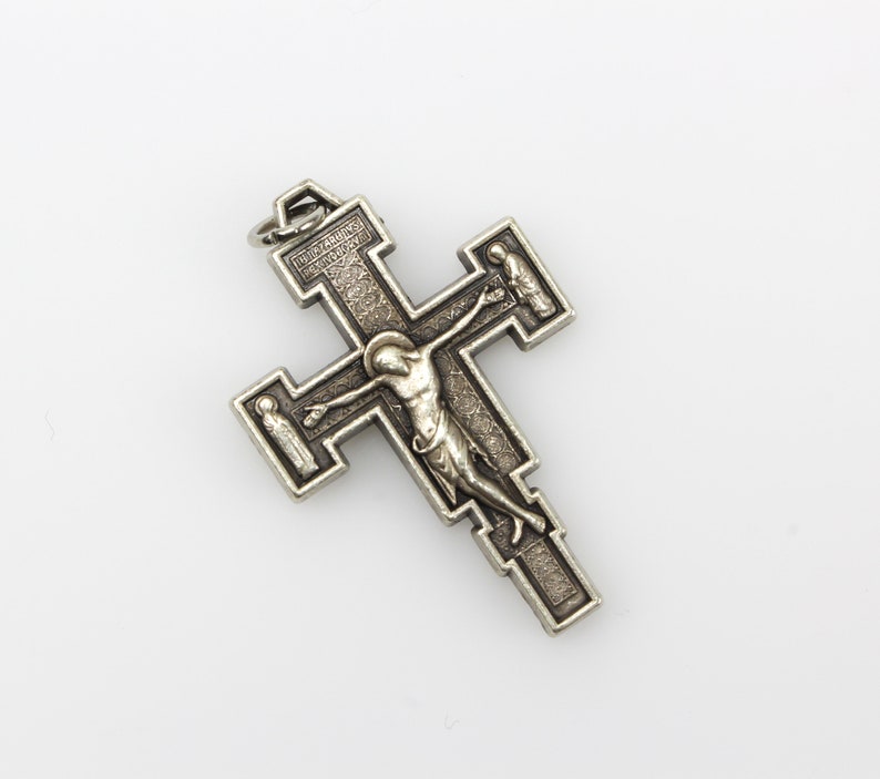 Op de afbeelding: Een zilveren metalen kruisbeeld met een gedetailleerd ontwerp. Het kruisbeeld toont een figuur van Jezus Christus aan het kruis, met twee kleinere figuren aan weerszijden. De inscriptie "INRI" is zichtbaar op het kruis.