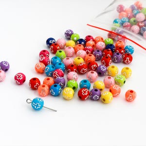 May include: A collection of colorful round beads in various shades, including red, blue, yellow, and green. Each bead features a cross design. Some beads are loose, while others are in a clear plastic bag. A single bead with a wire loop is also present.