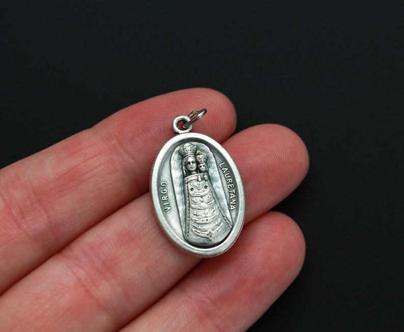 May include: Silver oval pendant with a depiction of the Virgin Mary and child. The pendant has the words "VIRGO LAURETANA" inscribed on it.