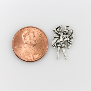 May include: Silver charm of an angel with wings outstretched, holding a sword. The charm is about the size of a US penny.