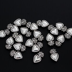 May include: A collection of silver-toned heart-shaped charms with a flame design. The charms are scattered on a black background.